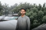 A young South Asian man stands outdoors with lush greenery in the background, exuding calmness.